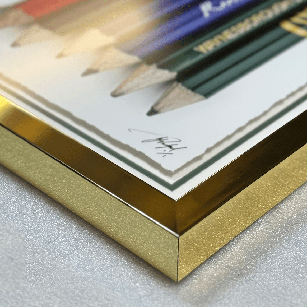 A close-up of a square picture frame corner with a metallic gold glitter finish, holding a white print featuring colored pencils, set on a textured light gray surface.